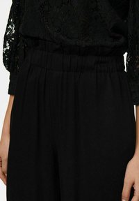 Zwarte kanten blouse met lange mouwen boven een geplooide, hooggetailleerde zwarte broek. De stof lijkt zacht te zijn met een gladde textuur, zonder zichtbare patronen.