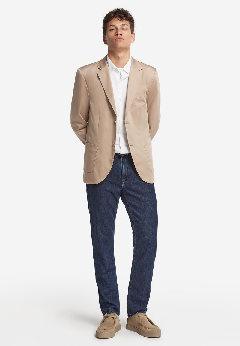 Beige Blazer mit Reverskragen, weißem Hemd, dunkelblauen Jeans und beigen Schuhen, stehend mit Händen hinter dem Rücken vor einem weißen Hintergrund.