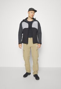 Giacca zip-up nera e grigia con cappuccio, pantaloni cargo beige e scarpe nere. La giacca presenta un design bicolore e un logo riflettente.