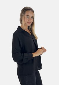 Zwarte blouse met lange mouwen, een V-hals en manchetten, gemaakt van een lichtgewicht, gladde stof; met een losse pasvorm en minimale details.