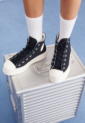 Füße in schwarzen High-Top-Sneakers mit weißen Sohlen und weißen Socken, die auf einer silbernen Metallbox stehen, vor einem blauen Bodenhintergrund.