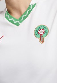 Jersey deportivo blanco con cuello en zigzag verde y el emblema de la Federación Real Marroquí de Fútbol en el pecho.