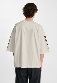 Persoon met krullend haar die wegkijkt, gekleed in een oversized lichtbeige T-shirt met zwarte diagonale strepen op beide mouwen en zwarte broek.