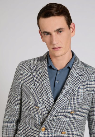 Joven de cabello castaño corto que lleva una camiseta polo azul bajo un blazer cruzado de estampado pata de gallo con botones dorados, sobre un fondo liso.