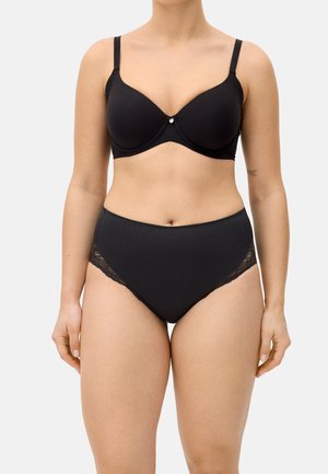 Mujer con sujetador negro con aros y bragas negras de cintura alta con ribete de encaje, de pie frente a un fondo claro y liso.