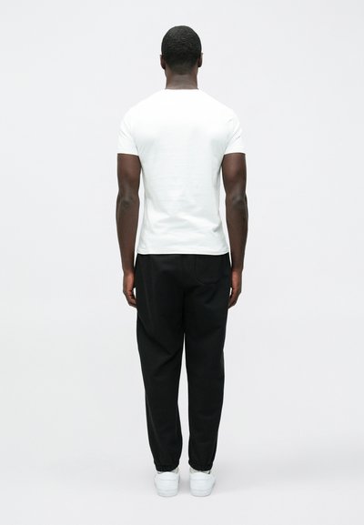 T-shirt branca de manga curta e calças de treino pretas, vista de costas. As calças têm cintura elástica; os sapatos são brancos.