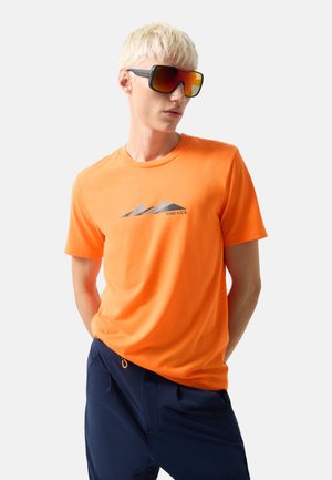 Man met kort blond haar die oversized oranje gradient zonnebril draagt, een oranje "FIRE+ICE" T-shirt en een donkerblauwe broek tegen een effen achtergrond.