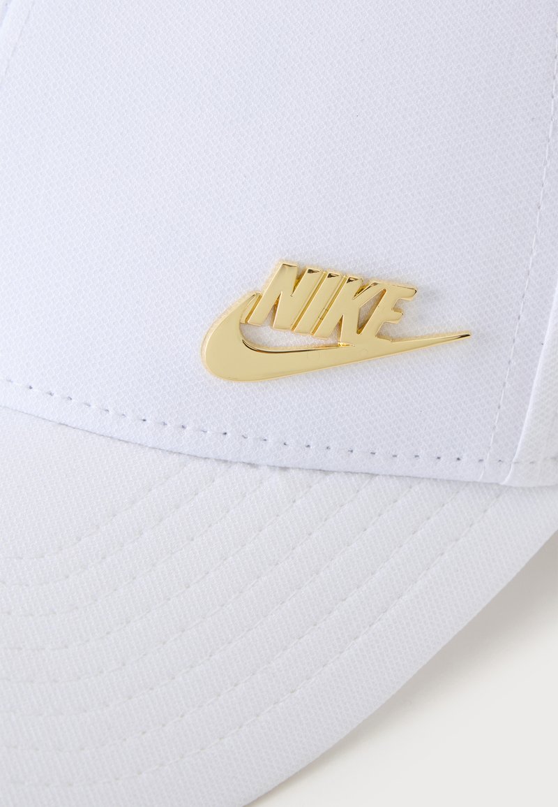 Casquette en tissu blanc avec un logo Nike métallique doré et un swoosh sur le panneau avant au-dessus de la visière.