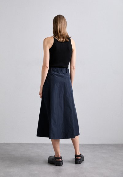 Jupe midi bleu marine avec une taille froncée, associée à un débardeur noir à côtes et des chaussures noires à enfiler, sur un fond neutre.