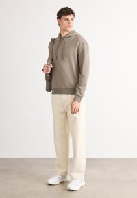 Brun hettegenser med snøring, kombinert med lysebeige bukser og hvite joggesko. Modellen holder en grå skulderveske. Enkelt design og passform.