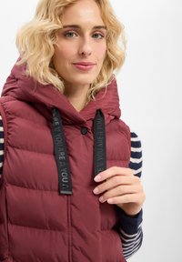 Weinrote Steppweste mit großer Kapuze, ausgestattet mit schwarzen Kordeln mit Text. Darunter ist ein gestreifter Pullover in Marineblau und Weiß sichtbar.