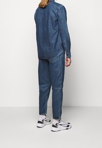 Camisa y pantalones de mezclilla de lavado oscuro, con un ajuste holgado y puños con botones. Zapatillas blancas y marinas completan el atuendo. Diseño simple y casual.