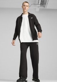 Sort træningsjakke med lynlås og Puma-logo, parret med sorte bukser. Outfittet har en hvid T-shirt under og sorte sneakers.