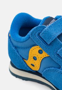 Blå og gul barne sneakers med borrelås, teksturert stoff og polstret krage, vist i nærbilde på hvit bakgrunn.