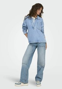 Un pulóver de denim azul claro con cuello y bolsillo en el pecho, combinado con unos jeans de tiro amplio azul claro que presentan detalles de parches en la parte frontal y un ajuste relajado.