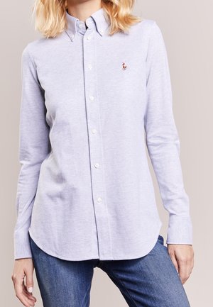 Femme portant une chemise bleu clair à boutons avec manches longues et petit logo brodé, associée à un jean bleu, sur fond neutre.