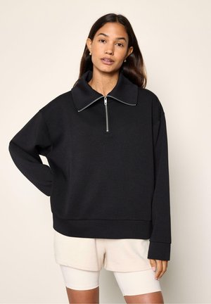 Donna che indossa un pullover nero a maniche lunghe con collo con cerniera frontale e pantaloncini beige, in piedi davanti a uno sfondo chiaro e uniforme.