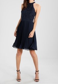 Navy blue dress with a high lace neck, fitted bodice, and pleated skirt. The fabric is soft with a flowing texture, complemented by straps and an A-line silhouette.
