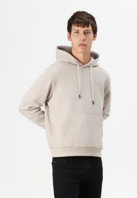 Beige Kapuzenpullover aus weichem Stoff, mit einer Fronttasche, verstellbarem Kapuzenzug und gerippten Bündchen, kombiniert mit schwarzen Jeans.