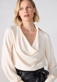 Kremfarget bluse med draperinger, lange ermer og myk krage, kombinert med et svart lærskjørt med knyttet midjebånd.