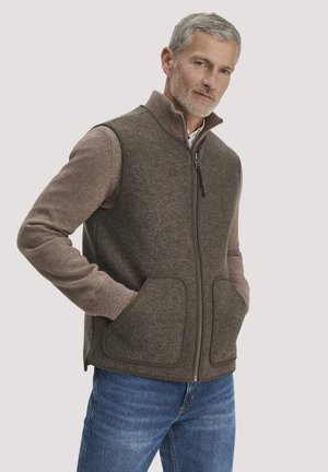 Mann mit grauem Haar und Bart trägt eine braune Fleeceweste mit Reißverschluss über einem langärmeligen Pullover und blauen Jeans, die Hände in den Westentaschen.