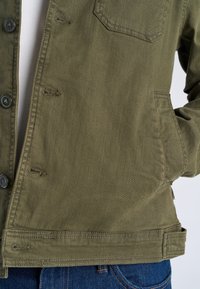 Person trägt eine olivgrüne, geknöpfte Jacke mit Taschen vorne und an der Brust, kombiniert mit blauen Jeans.