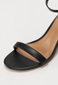 Sandale noire à talon haut ouverte à l’avant, avec une seule bride frontale et une bride à la cheville, avec une semelle intérieure beige et une texture en cuir lisse.