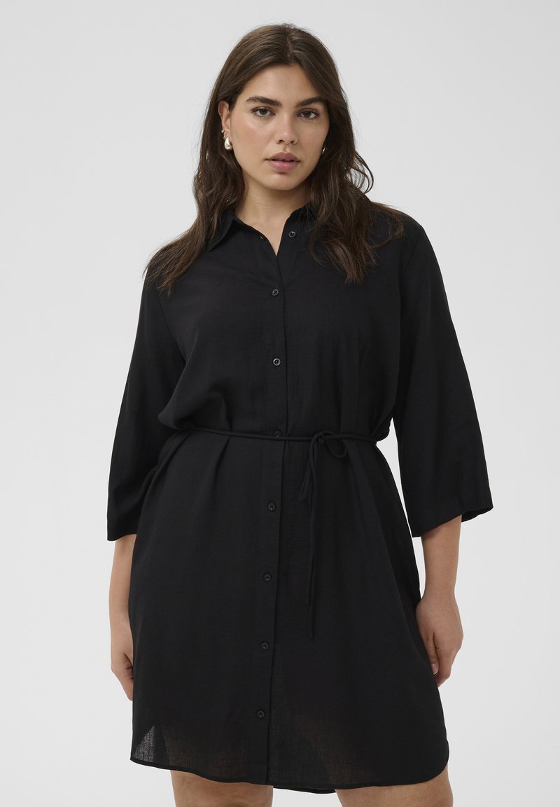 Femme portant une robe-chemise noire boutonnée avec des manches trois-quarts et une ceinture à la taille, debout devant un fond clair uni.