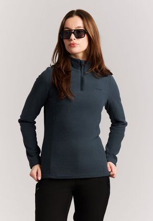 Frau in dunkelblauem Fleecepullover und schwarzen Hosen mit großen schwarzen Sonnenbrillen, stehend mit Händen an den Hüften vor einfarbigem Hintergrund.
