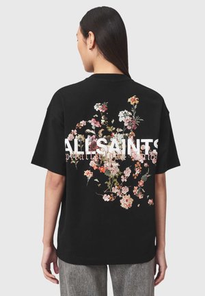 Kvinde iført sort oversized t-shirt med lyserødt og hvidt blomsterdesign og teksten "ALLSAINTS" på bagsiden, kombineret med grå bukser.