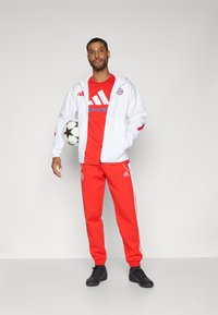 Red tracksuit with white accents, featuring a zip-up jacket and logoed t-shirt. Black athletic shoes. Player holds a soccer ball.