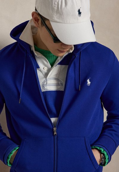 Homme portant une casquette blanche, des lunettes de soleil foncées, un sweat à capuche bleu zippé par-dessus un maillot de rugby vert et blanc rayé avec le col relevé, mains dans les poches.
