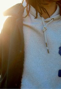 Person med dreadlocks som bär en ljusgrå hoodie och en mörk jacka i solskenet, med hoodie-snoddarna knutna i en lös knut.