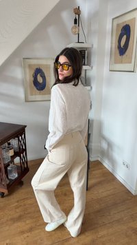 Femme en pantalon beige et blouse blanche à œillets, portant des lunettes de soleil teintées ambre, debout dans une pièce avec une œuvre d'art abstraite bleue encadrée et un sol en bois.