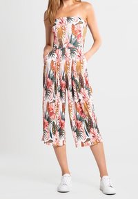 Combinaison fleurie sans bretelles blanche, ornée de fleurs roses et de feuillage vert, avec une coupe ample et courte et des poches latérales. Portée avec des baskets blanches.