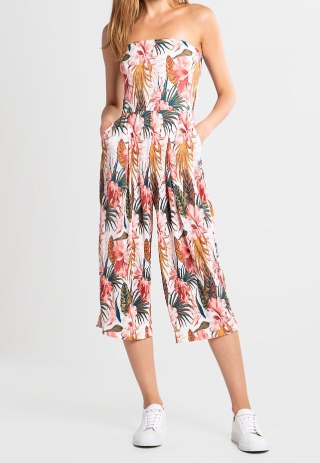 Combinaison fleurie sans bretelles blanche, ornée de fleurs roses et de feuillage vert, avec une coupe ample et courte et des poches latérales. Portée avec des baskets blanches.