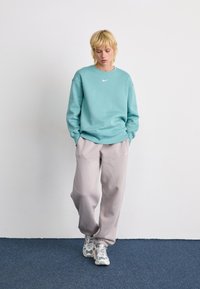 Blond person står mod en hvid væg iført en teal sweatshirt, løse beige bukser og hvide-grønne sneakers med hænderne i lommen.