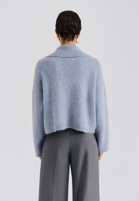 Πουλόβερ από μαλλί μοχέρ ανοιχτού μπλε χρώματος με κοντό κόψιμο, φαρδιές μανσέτες και διπλωμένο γιακά, συνδυασμένο με γκρι ραμμένο παντελόνι. Απαλή, υφή με υφασμάτινο φινίρισμα.