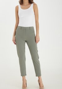 Kvinna i en figursydd vit ärmlös topp, olivgröna skräddarsydda anklebyxor och beiga högklackade skor som står framför en vit bakgrund.