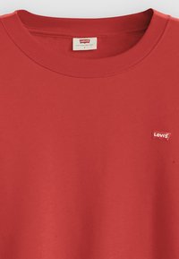 T-shirt en coton rouge avec un col rond. Présente un petit patch logo Levi's rouge et blanc sur la poitrine gauche. Texture lisse.