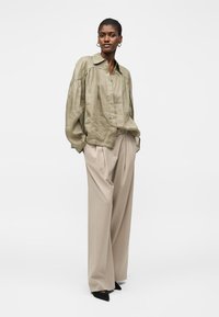 Γυναίκα φοράει φαρδιά πουκάμισα σε ελιάς και πλατύχωρα μπεζ παντελόνια, στέκεται με τα χέρια στις τσέπες και μαύρα μυτερά παπούτσια.