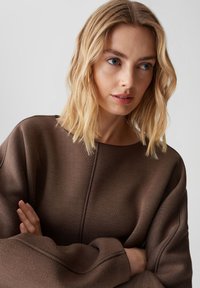 Maglione oversized marrone con scollo rotondo, tessuto testurizzato e cuciture sottili; modella in posa con le braccia incrociate, acconciata con onde morbide.