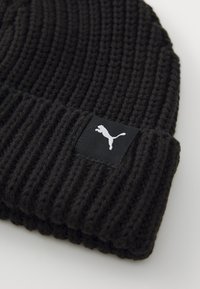 Căciulă neagră din tricot cu textură canelată, margine pliată și un eticheta neagră cu un logo alb Puma pe partea din față.
