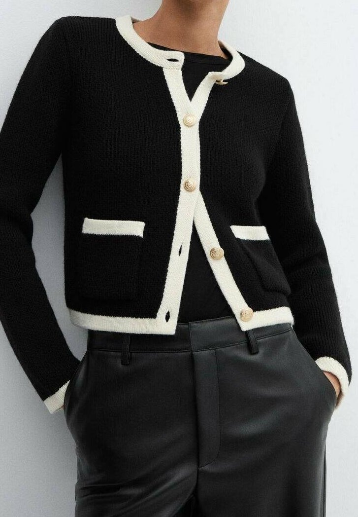 Cardigan en maille noire avec bordure blanche, boutons dorés, deux poches avant, porté sur un top noir et associé à un pantalon en cuir noir.