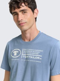 Vaaleansininen puuvillainen t-paita pyöreällä pääntiellä. Edessä valkoinen painettu teksti ja pyöreä logo. Lyhyet hihat, rento istuvuus.