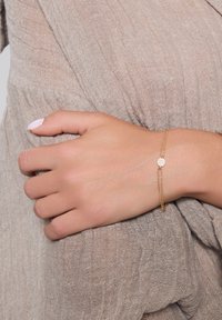 Złota bransoletka z delikatnym łańcuszkiem, z małym okrągłym zawieszką wysadzanym białymi kamieniami, noszona na opalonej ręce na tle teksturowanej tkaniny.