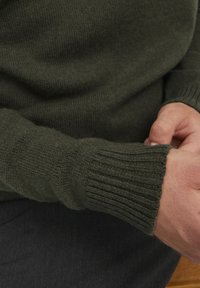Pull en laine tricotée de couleur vert foncé avec des poignets côtelés. Texture douce, coupe ajustée et finition unie, porté sur un pantalon sombre.