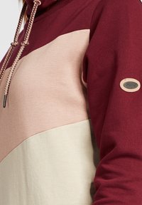 Sweat à capuche bicolore bordeaux, rose clair et crème avec cordons tressés et petite étiquette de marque "OXMO" sur la manche.