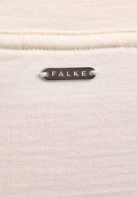 Kremowa tkanina o delikatnej fakturze prążków; zawiera srebrną metkę z napisem "FALKE" przymocowaną białą nicią.