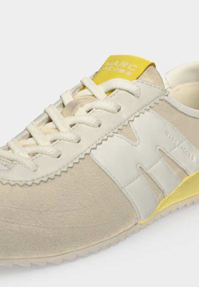 Baskets beige et blanches avec lacets blancs, talon et languette jaunes, arborant le logo Marc Jacobs sur la languette et le côté.